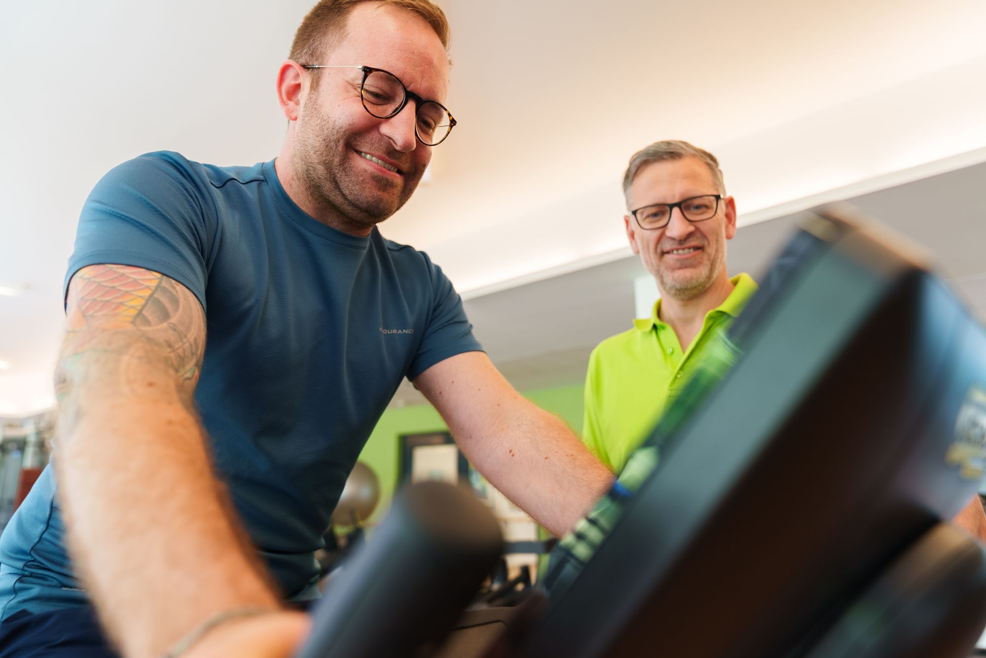 Therapeut des Therapie- & Gesundheitszentrums Dentl betreut einen lachenden Patienten am Trainingsgerät